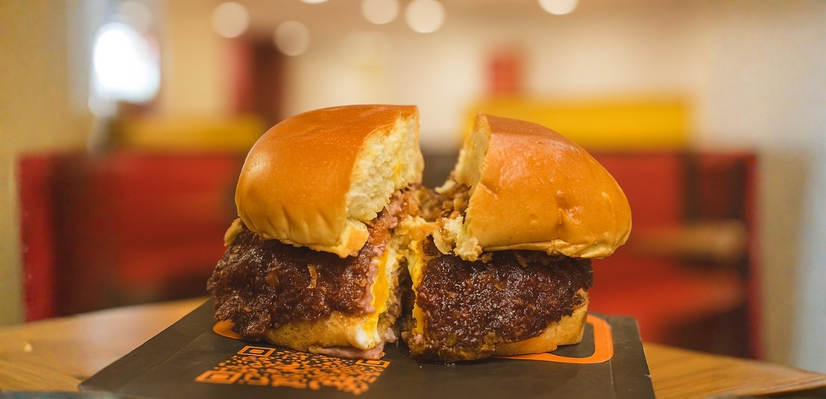Hamburguesa cortada por la mitad con pan brioche, queso fundido y bordes crujientes de carne, sobre soporte negro con diseño en interior de Yogui Burger (Torrejón de Ardoz).